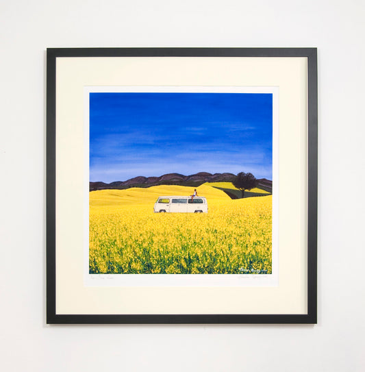 Beautifully framed high quality giclee print of free roam, a man sitting on the roof of a white campervan on a sunny day of blue skies in yellow flower fields with trees and hills in the background. Contemporary high quality affordable art prints.